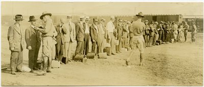 Fotografía horizontal en sepia que muestra un grupo de hombres en ropa civil, muchos de los cuales tienen equipaje, parados en una fila. Soldados uniformados caminan a lo largo de la fila hablando con los hombres individualmente. Se pueden ver vías de tren de Carl Michel